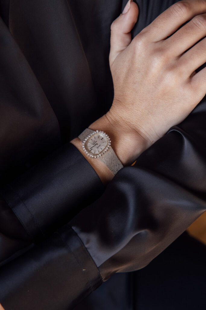 Reloj de señora con esfera plateada, bisel engastado con piedras blancas y brazalete de metal blanco texturizado, en una muñeca sobre satén negro