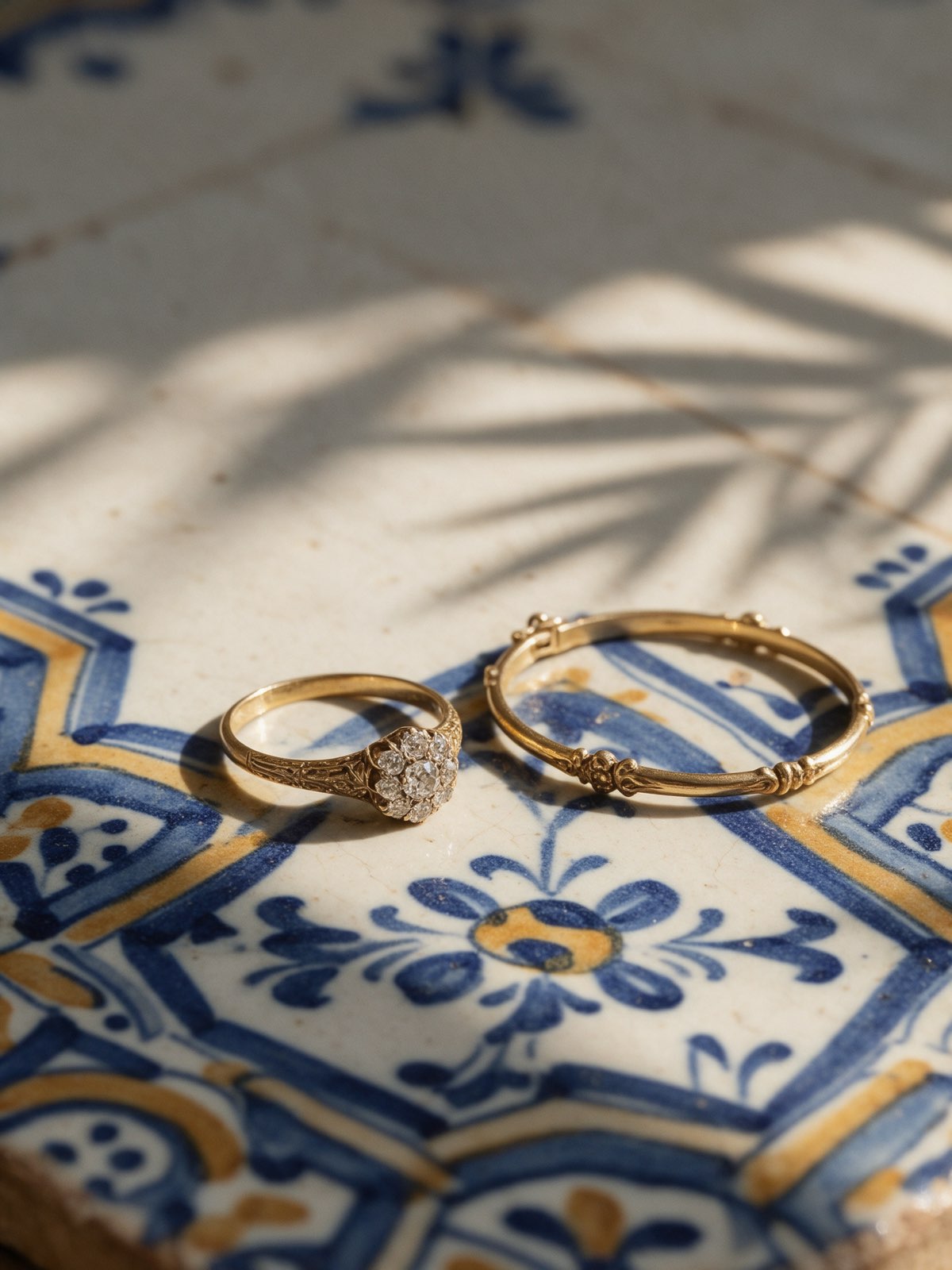 Bodegón de una sortija y una pulsera de oro sobre un azulejo andaluz pintado a mano en azul y ocre, con la sombra de unas palmeras a la luz de la mañana
